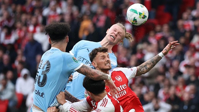 Soccer players in red-and-white and light-blue kits jump for a header as the ball rises above them.