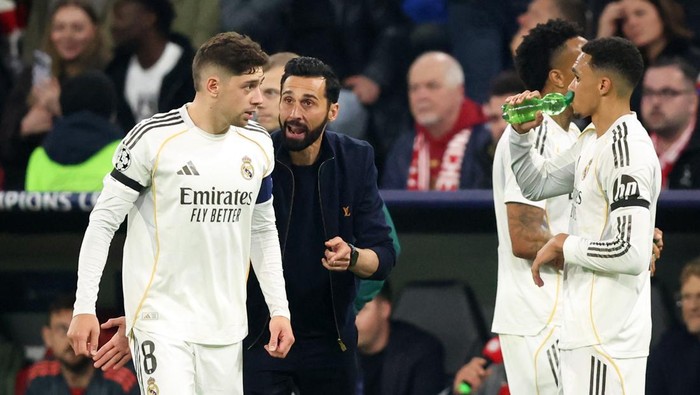 Real Madrid players on the pitch as coach Zinedine Zidane talks to the player on the left; another teammate drinks water on the right.
