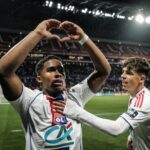 Two football players in white Lyon kits celebrate on the pitch; one forms a heart shape with his arms as a teammate pats him on the chest in front of a cheering stadium crowd.