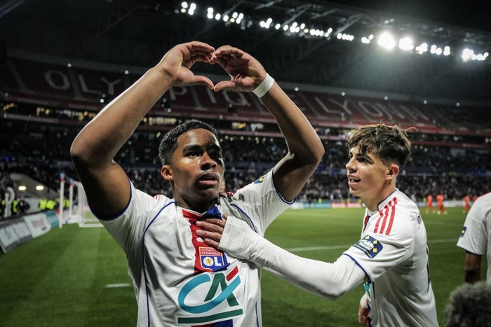 Two football players in white Lyon kits celebrate on the pitch; one forms a heart shape with his arms as a teammate pats him on the chest in front of a cheering stadium crowd.