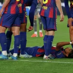 Soccer players gather around an injured teammate lying on the grass, one player lying on his back with hands near his face while others stand nearby in blue and maroon uniforms.