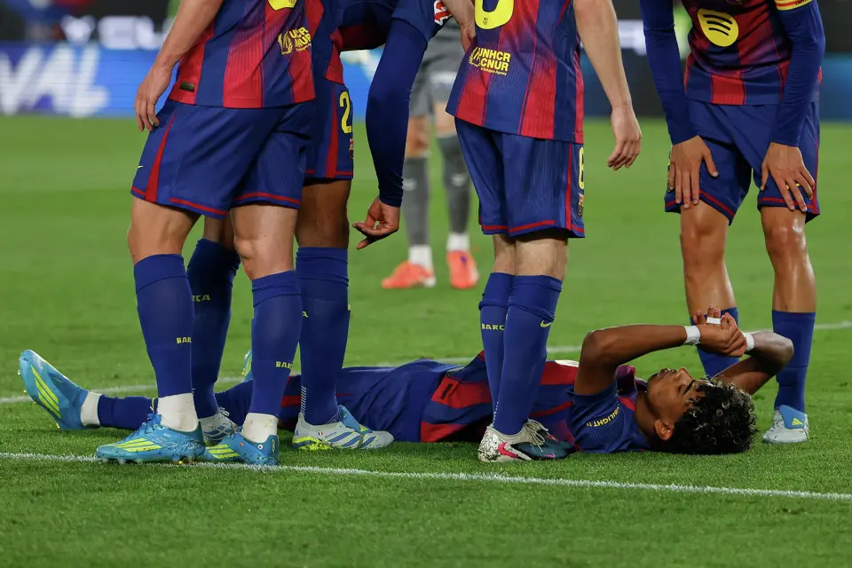 Soccer players gather around an injured teammate lying on the grass, one player lying on his back with hands near his face while others stand nearby in blue and maroon uniforms.