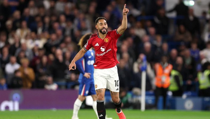 Manchester United player in red kit pointing upward after a goal on a football pitch with fans in the stands behind him