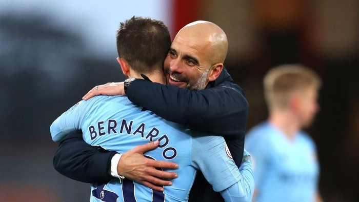 Two men in light blue football kits hug on the pitch, with Bernardo Silva's name visible on one jersey.