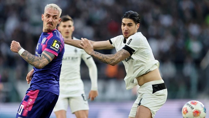 Two football players clash for the ball during a match; one in a purple kit, the other in white, with a ball nearby and spectators in the stands behind them.