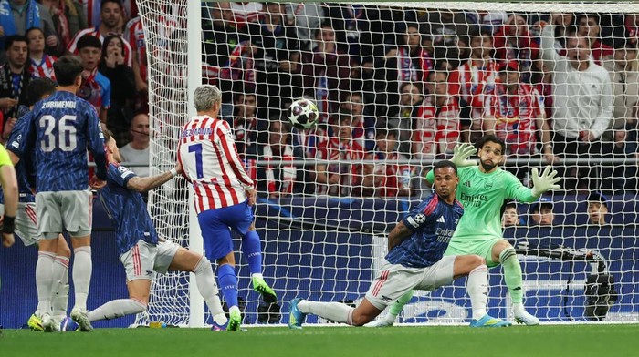 Soccer action near the goal: striker in red-and-white stripes shoots as the ball heads toward the net, goalkeeper in green attempts a save with teammates nearby.