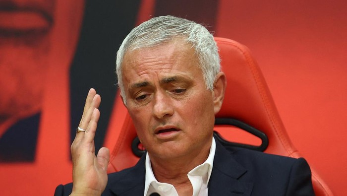 Man with gray hair in a dark suit sits in a bright red seat, raising his left hand in a questioning gesture.