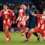 Soccer match action with Bayern players in red chasing the ball against a dark-clad opponent on a green field.