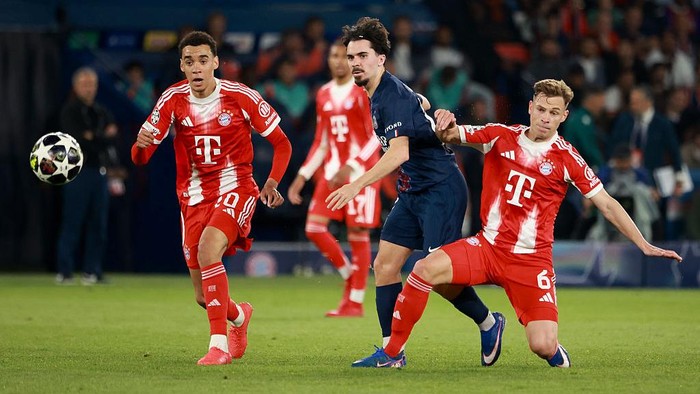 Soccer match action with Bayern players in red chasing the ball against a dark-clad opponent on a green field.