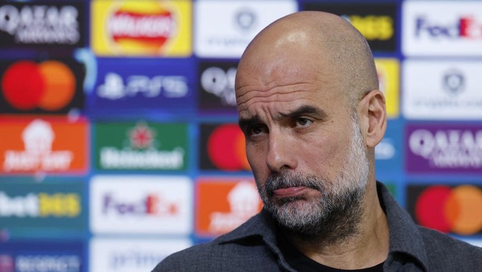 Bearded, bald man in a dark shirt looks to the side during a press conference with sponsor logos in the background.