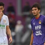 Two soccer players on a field during a match: left player in a white kit touches his nose, right player in a purple kit looks ahead.