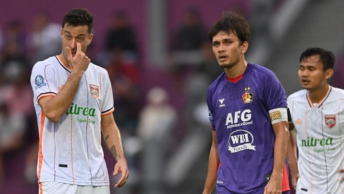 Two soccer players on a field during a match: left player in a white kit touches his nose, right player in a purple kit looks ahead.