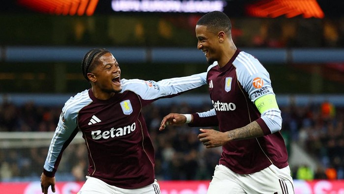 Two Aston Villa teammates celebrate a goal on the pitch, one with an arm around the other as they smile broadly on the field.