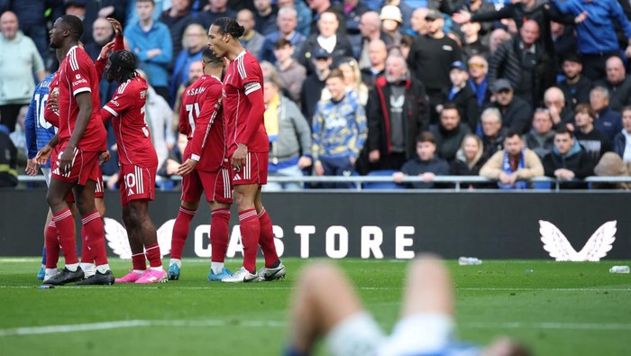 Soccer players in red uniforms walk together on a green field as spectators watch from the stands behind them, with a blurred person lying in the foreground.