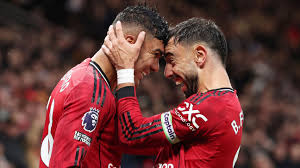 Two soccer players in red jerseys celebrate a goal with a close forehead-to-forehead hug on the field.