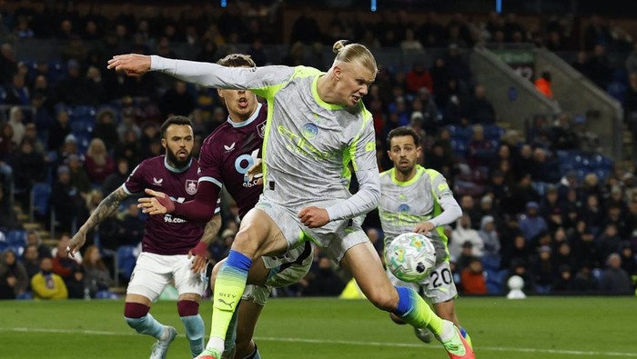 Soccer player in a gray kit kicks the ball while pursuing rivals on a night-time stadium field.