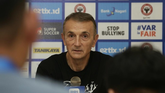 A man speaks at a press conference, sits behind a microphone with a backdrop featuring logos and a 'STOP BULLYING' sign in the background.