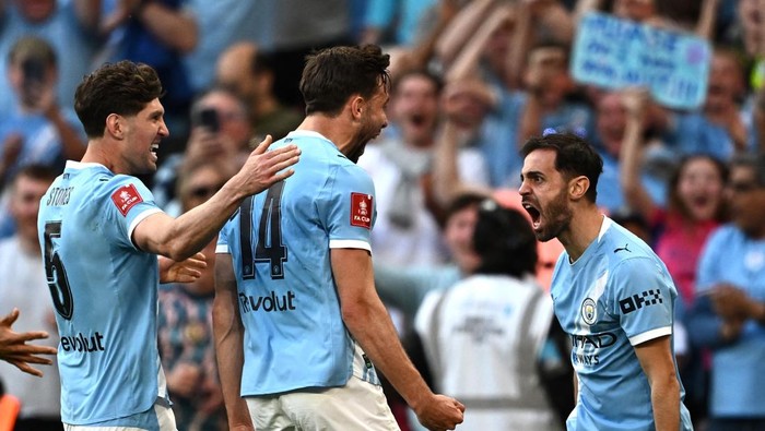 Three Manchester City players celebrate a goal on the pitch with fans cheering in the background.