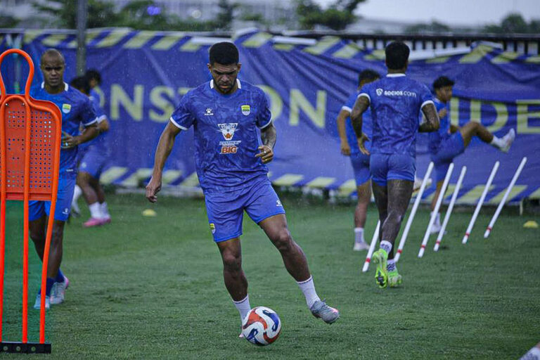Soccer players in blue training kits dribbling a ball on a grass field during a drill with cones nearby.
