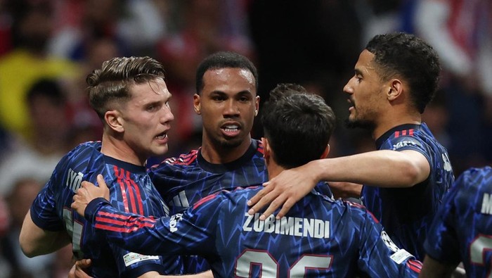 Group of blue-clad soccer players celebrate by embracing on the field after a play; one jersey reads Zubimendi 25.