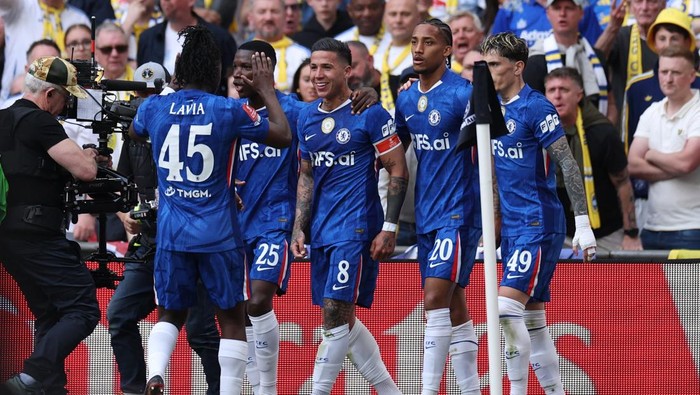 Soccer players in blue jerseys celebrate together on the field as a cameraman films the moment, crowd in the background.
