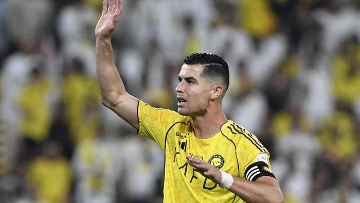Professional soccer player in a yellow jersey raises his right hand and waves to the crowd, wearing a captain's armband in a stadium crowd.