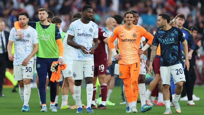 Soccer players from a team in white and orange kits walk together on the pitch after a match, some with orange vests in the background
