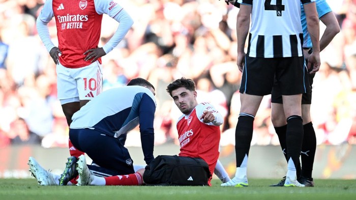 Arsenal player in red jersey sitting on the grass with a staff member leaning in, appears injured, as teammates and opponents in blue and black-and-white striped kits stand nearby on a soccer field with a crowded stadium background.