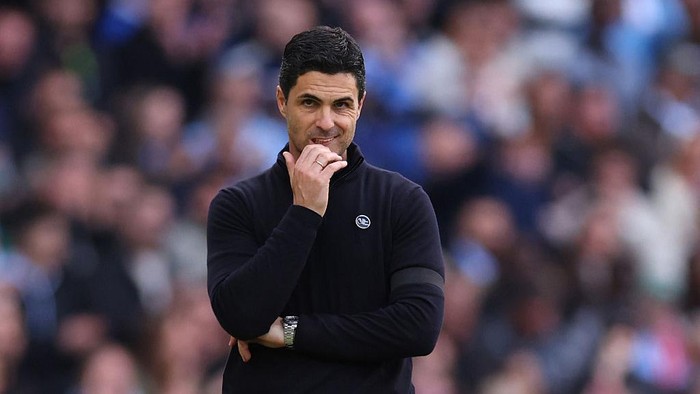 Man wearing a dark sweater on a soccer field, hand resting on chin in a thoughtful pose, blurred crowd behind him.