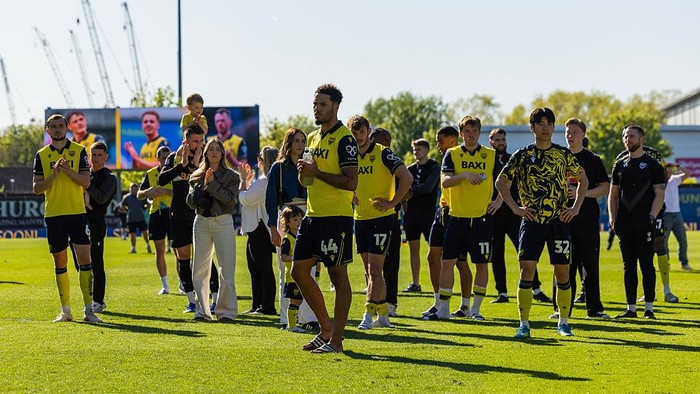 Soccer team in yellow and black uniforms standing on a sunny grass field with spectators in the background.