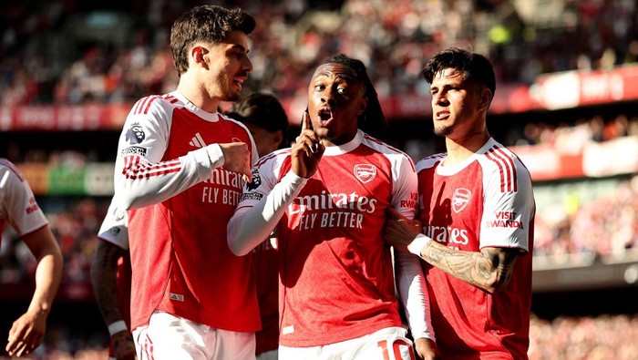 Three Arsenal players celebrate a goal on the pitch in red-and-white kits. The center player gestures with his finger while teammates commemorate the moment nearby.