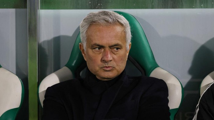 Football manager sitting on a green and white dugout bench with a stern expression.