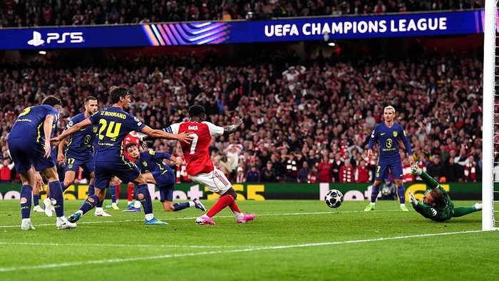 UEFA Champions League action: a red-shirted striker shoots toward goal while defenders challenge and the goalkeeper dives.