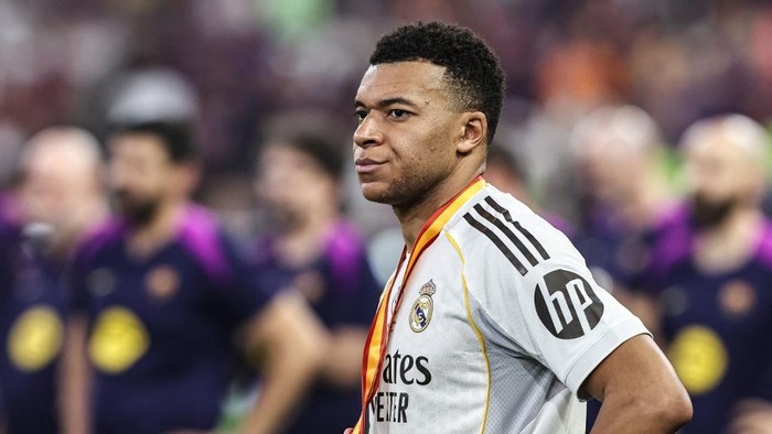 Footballer in a Real Madrid kit wearing a gold medal around his neck on the pitch after a match, with blurred teammates in the background.