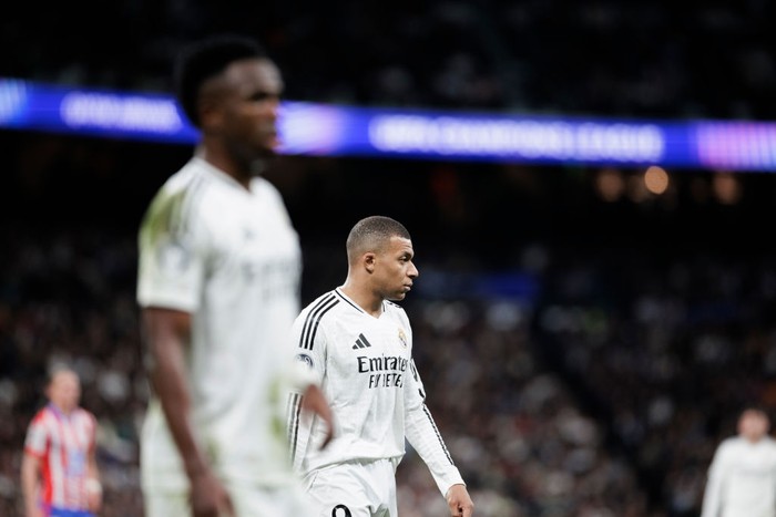 Real Madrid footballer in a white kit walking on the pitch during a match, with a teammate out of focus in the foreground and a blurred stadium crowd in the background.