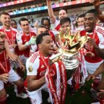 Arsenal players celebrate winning the Premier League trophy on the pitch, wearing red and white jerseys with medals.