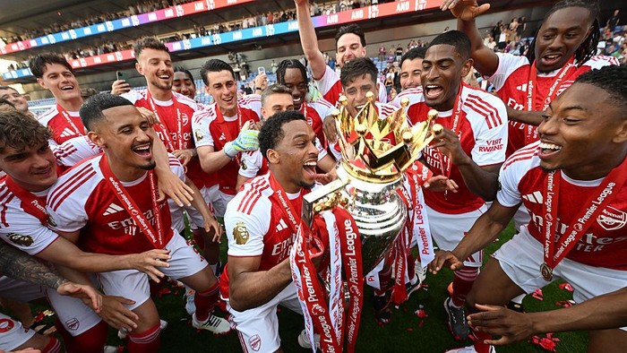 Arsenal players celebrate winning the Premier League trophy on the pitch, wearing red and white jerseys with medals.