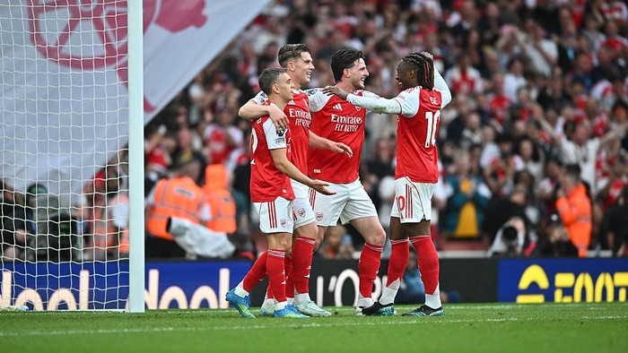 Arsenal players in red and white kits celebrate a goal on the pitch near the goalpost with cheering fans in the background.