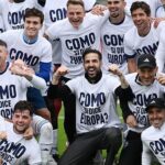 Group of soccer players celebrating together on a field, wearing white shirts with bold text on them.