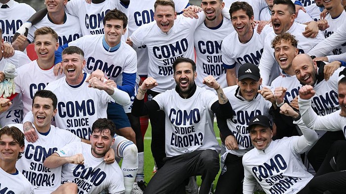 Group of soccer players celebrating together on a field, wearing white shirts with bold text on them.