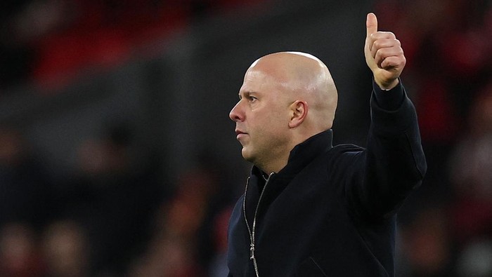Bald man in a dark jacket giving a thumbs-up gesture in a stadium.