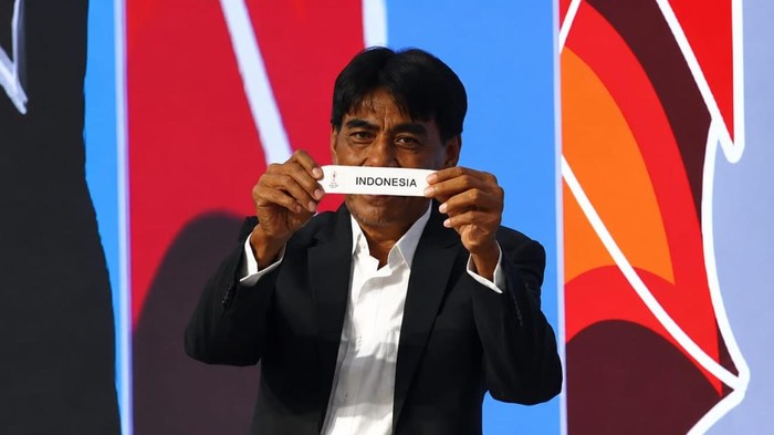 Man in a dark suit holds a white strip reading 'INDONESIA' in front of a colorful backdrop.