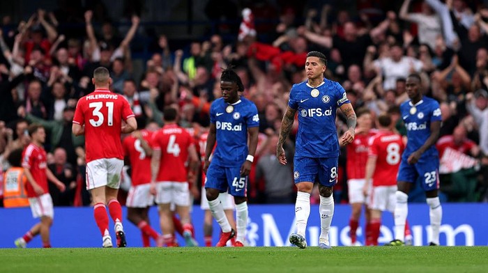 Soccer match on a green field with blue-jersey players in the foreground and red-jersey opponents celebrating in the background, stadium full of fans.