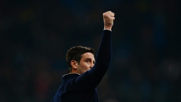 Man in a dark jacket raises a clenched fist in celebration, with a blurred stadium crowd in the background.