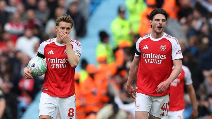Two Arsenal players in red kits walk on a football pitch, one holding a ball as fans watch from the stands behind them.