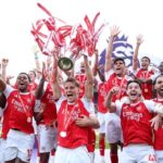 Arsenal FC players celebrate winning a trophy on the pitch, lifting a large trophy amid cheering teammates.