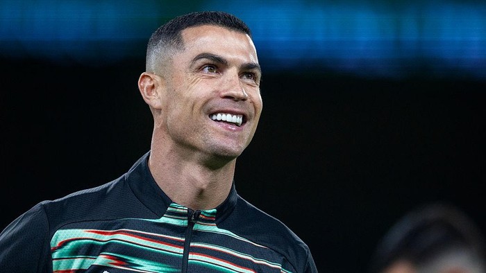Smiling man with short dark hair in a dark jacket with green and red stripes, against a dark background.