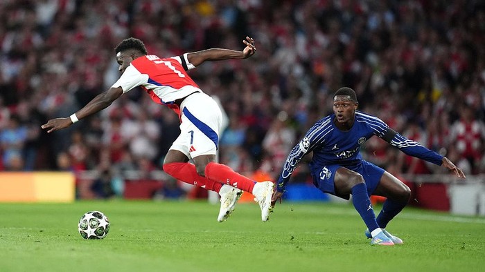 Two soccer players in red/white and blue kits compete for the ball on a green field during a match.