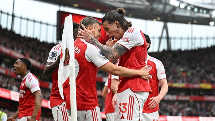 Arsenal players celebrate a goal, embracing near the corner flag on the pitch.