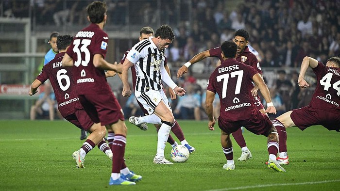 Soccer player in a white and black striped kit dribbles the ball while maroon-clad opponents converge on a green pitch.